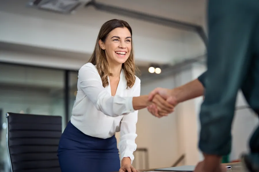 Professional woman in white blouse and navy skirt smiling and shaking hands in office setting during meeting.