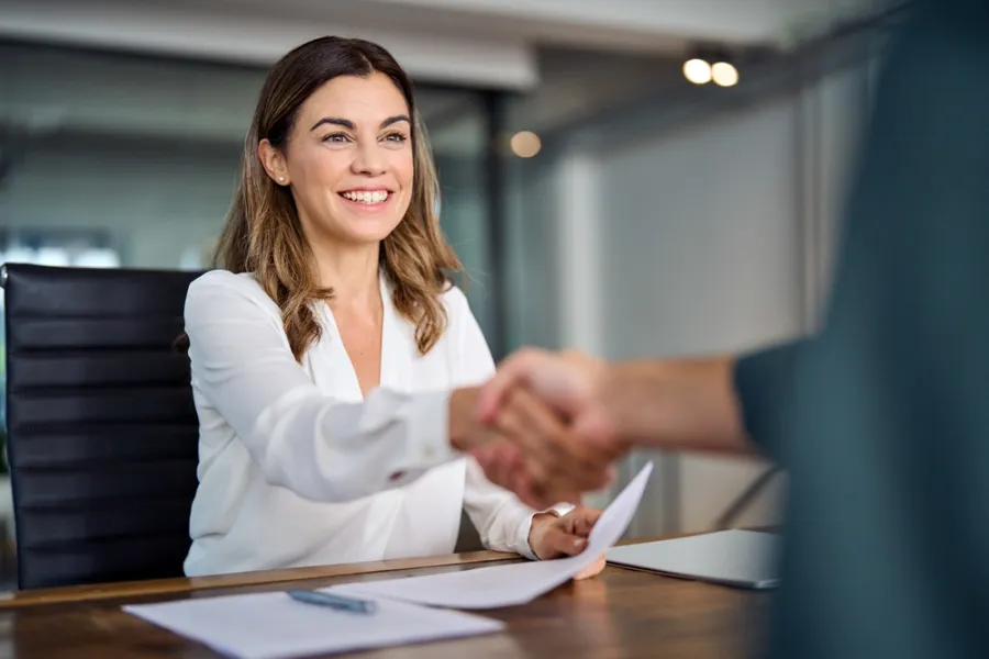 Smiling businesswoman shaking hands during a meeting holding papers at a modern office desk.