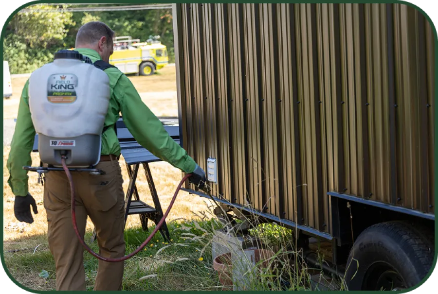 Man wearing green shirt and brown pants sprays herbicide near a metal building on a sunny day.