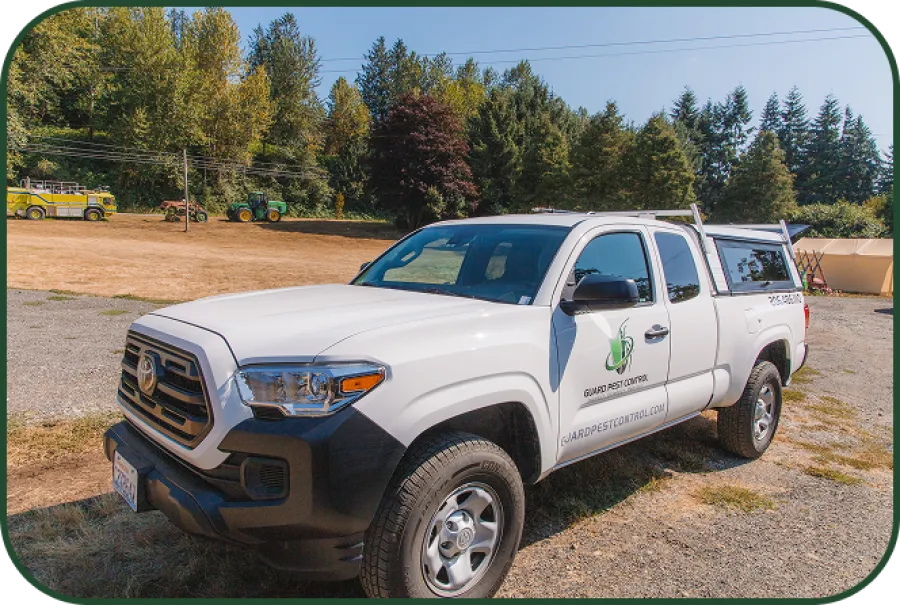 White Toyota Tacoma pickup truck with Guard Pest Control logo parked on gravel near trees and farm equipment.