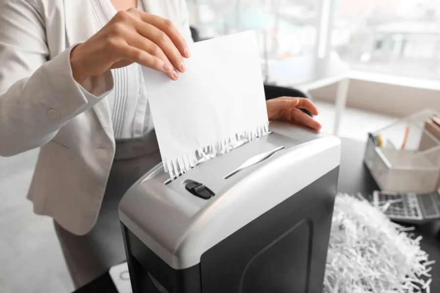 Person shredding documents with a paper shredder in an office with a pile of shredded paper nearby