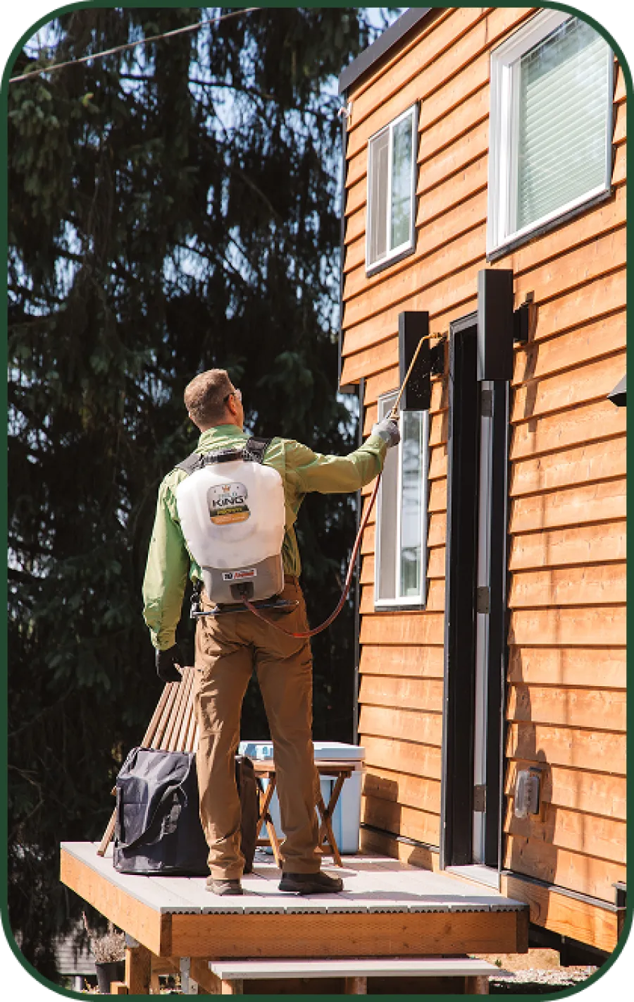 Technician spraying pest control treatment on wooden house exterior using a backpack sprayer on a sunny day