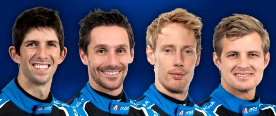 Four male race car drivers wearing blue and black racing suits, smiling against a blue background.