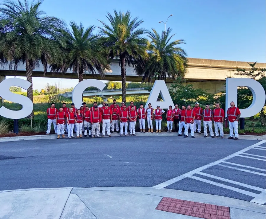 Group of people in red shirts and white pants standing in front of large SCAD letters with palm trees and overpass.