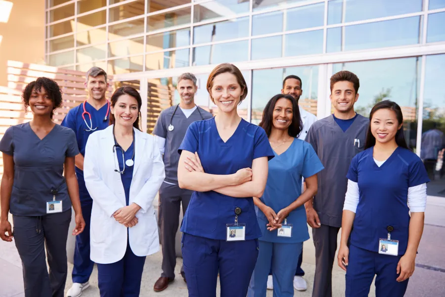 Diverse group of smiling healthcare professionals standing outside a medical facility in scrubs and lab coats.