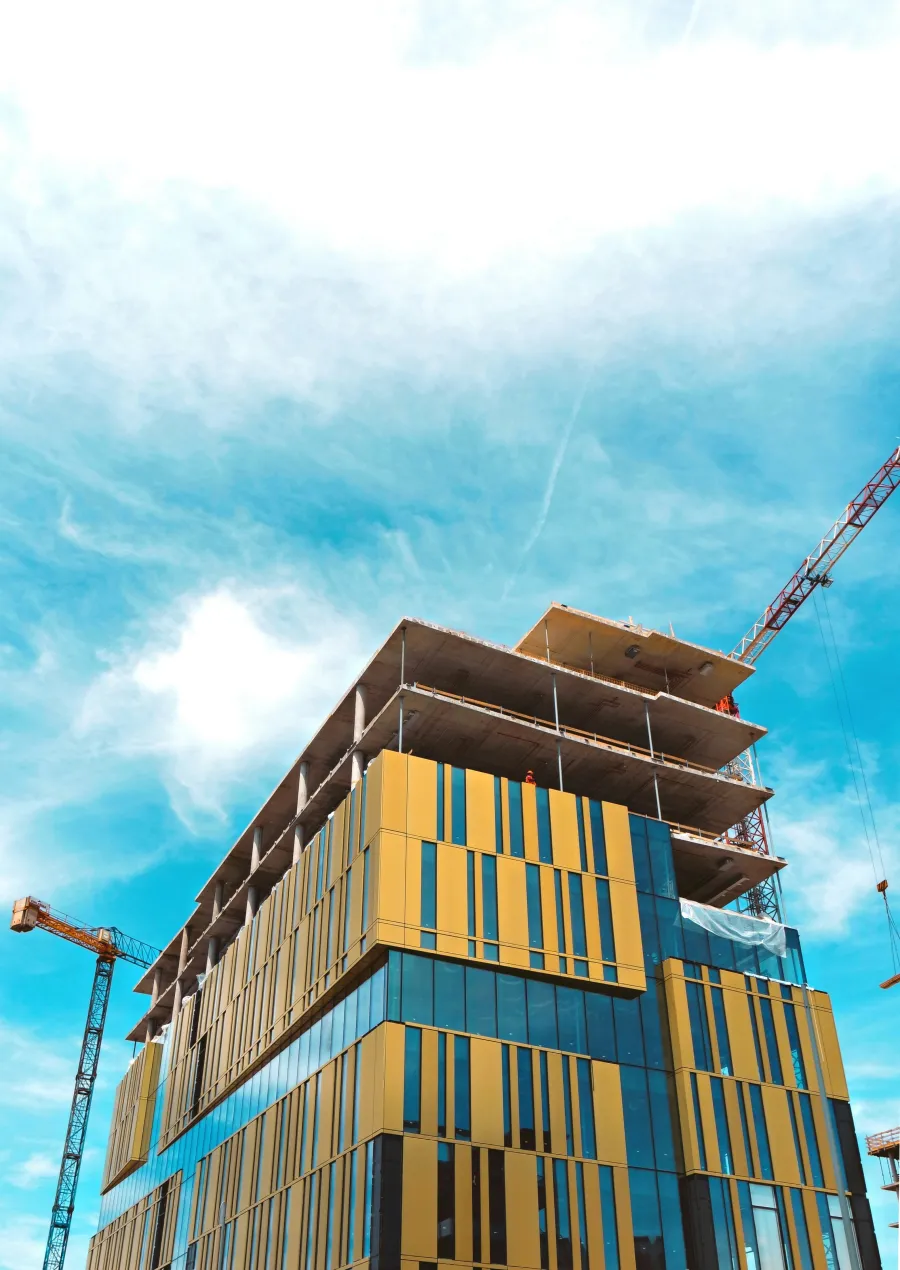 Modern building under construction with yellow and glass panels and cranes against a blue sky.