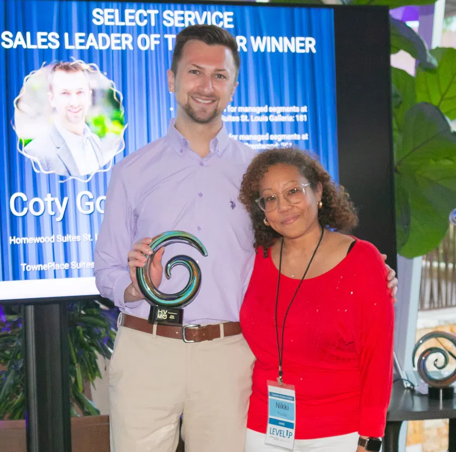 Smiling man holding a spiral award with a woman in red beside him at a sales leader recognition event.