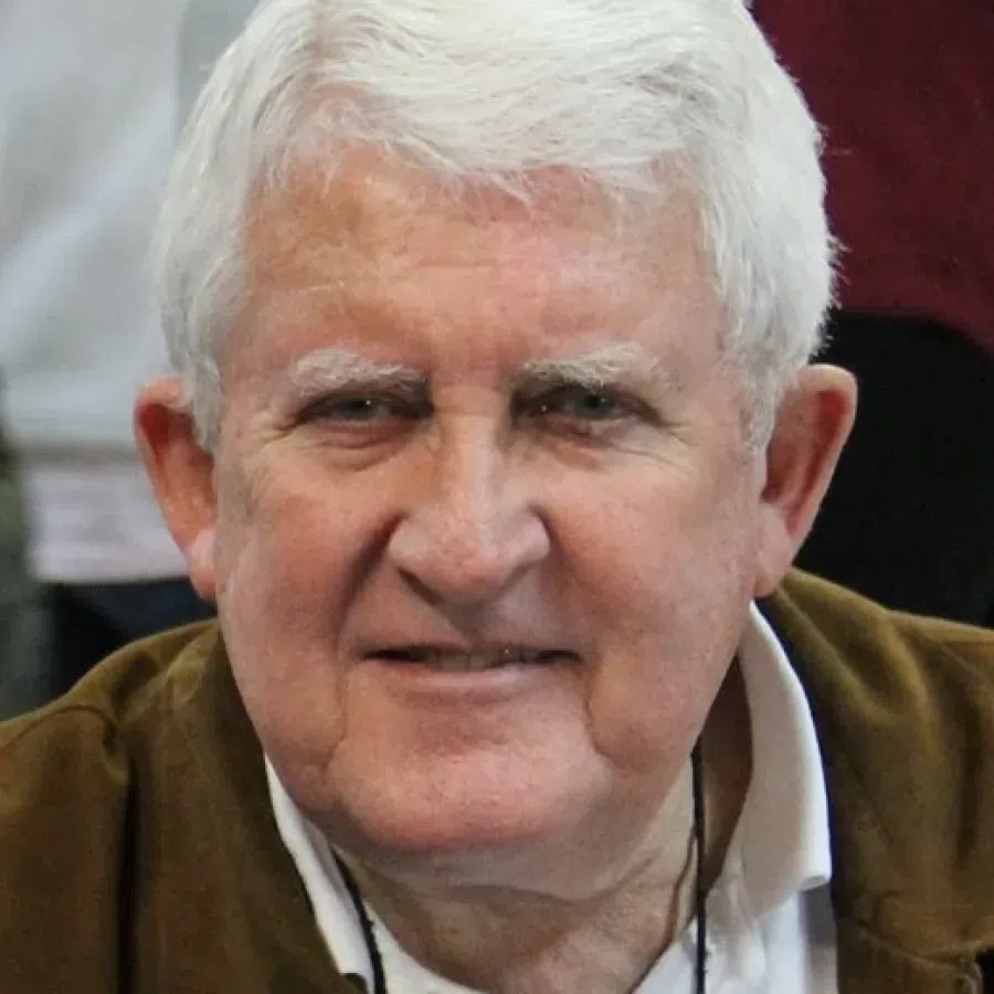 Close-up of a smiling elderly man with white hair wearing a brown jacket and white shirt.