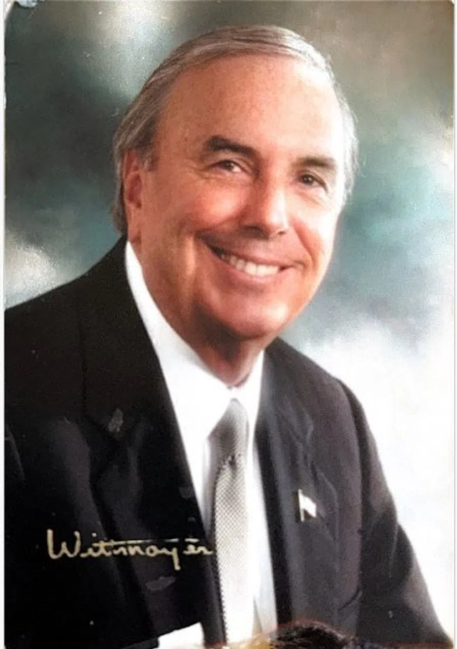 Smiling older man in a black suit and tie with a background of soft, blurred colors.