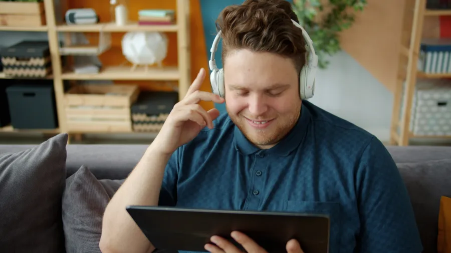 Smiling young man wearing headphones and using a tablet while sitting on a couch at home.