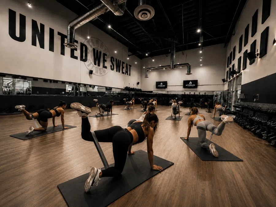 Group fitness class in modern gym performing leg lifts on yoga mats with slogan United We Sweat on wall