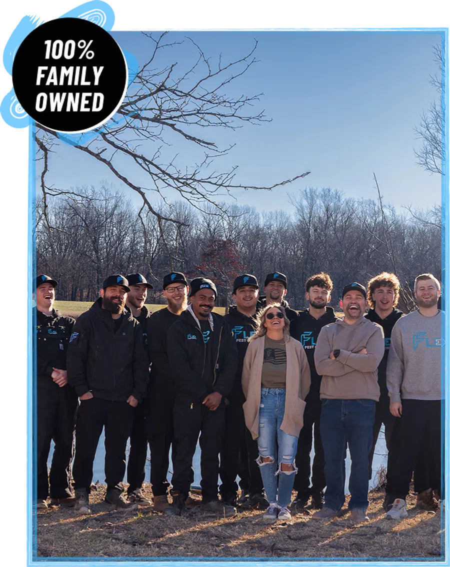 Group of casually dressed people smiling outdoors near a lake with leafless trees in winter under clear blue sky.