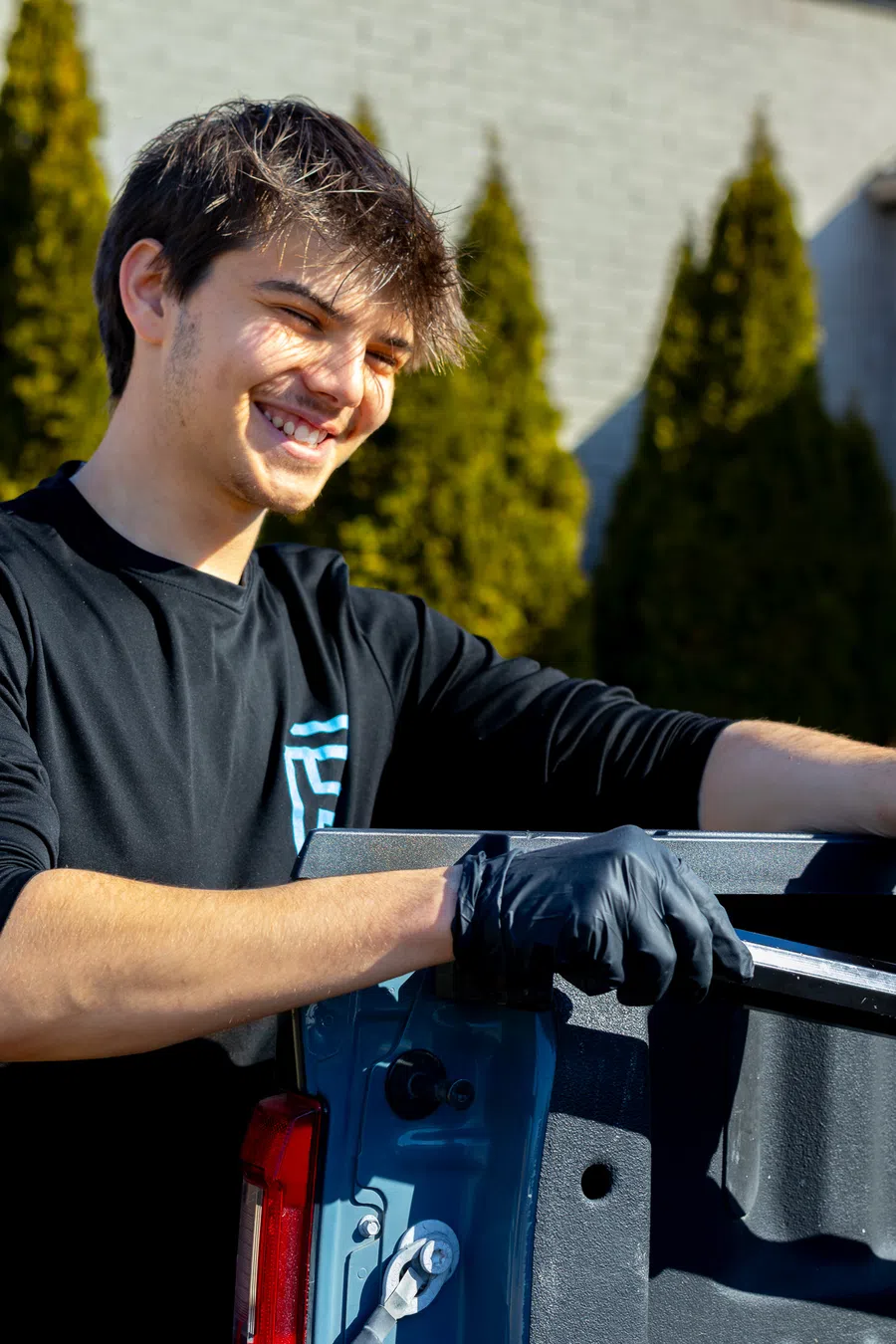 Young man wearing black gloves smiling while working on a blue vehicle outdoors with blurred trees in background