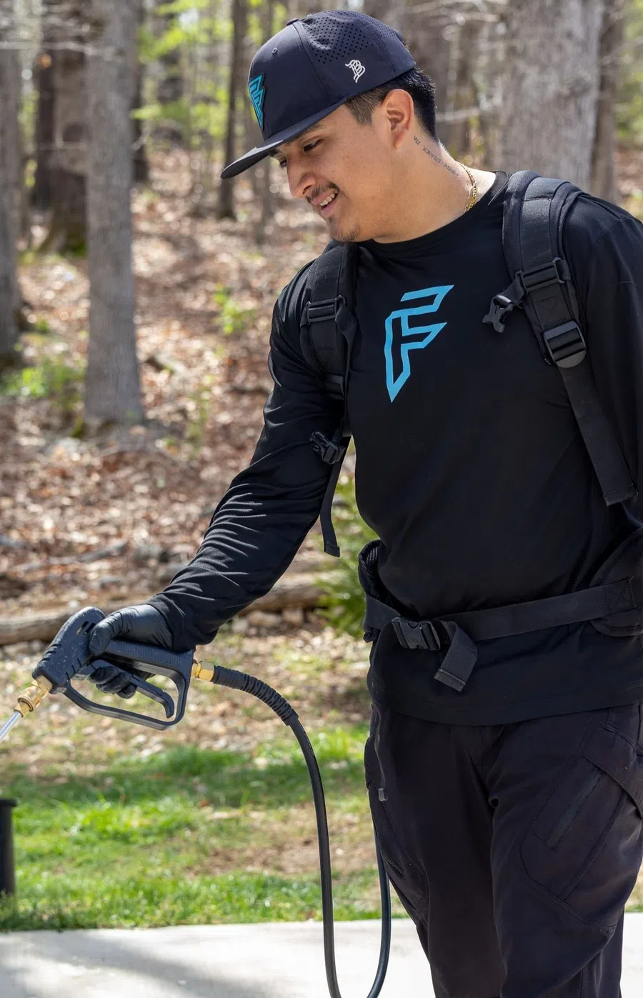 Man wearing black outfit and hat using pressure washer outdoors in a wooded area during daytime.
