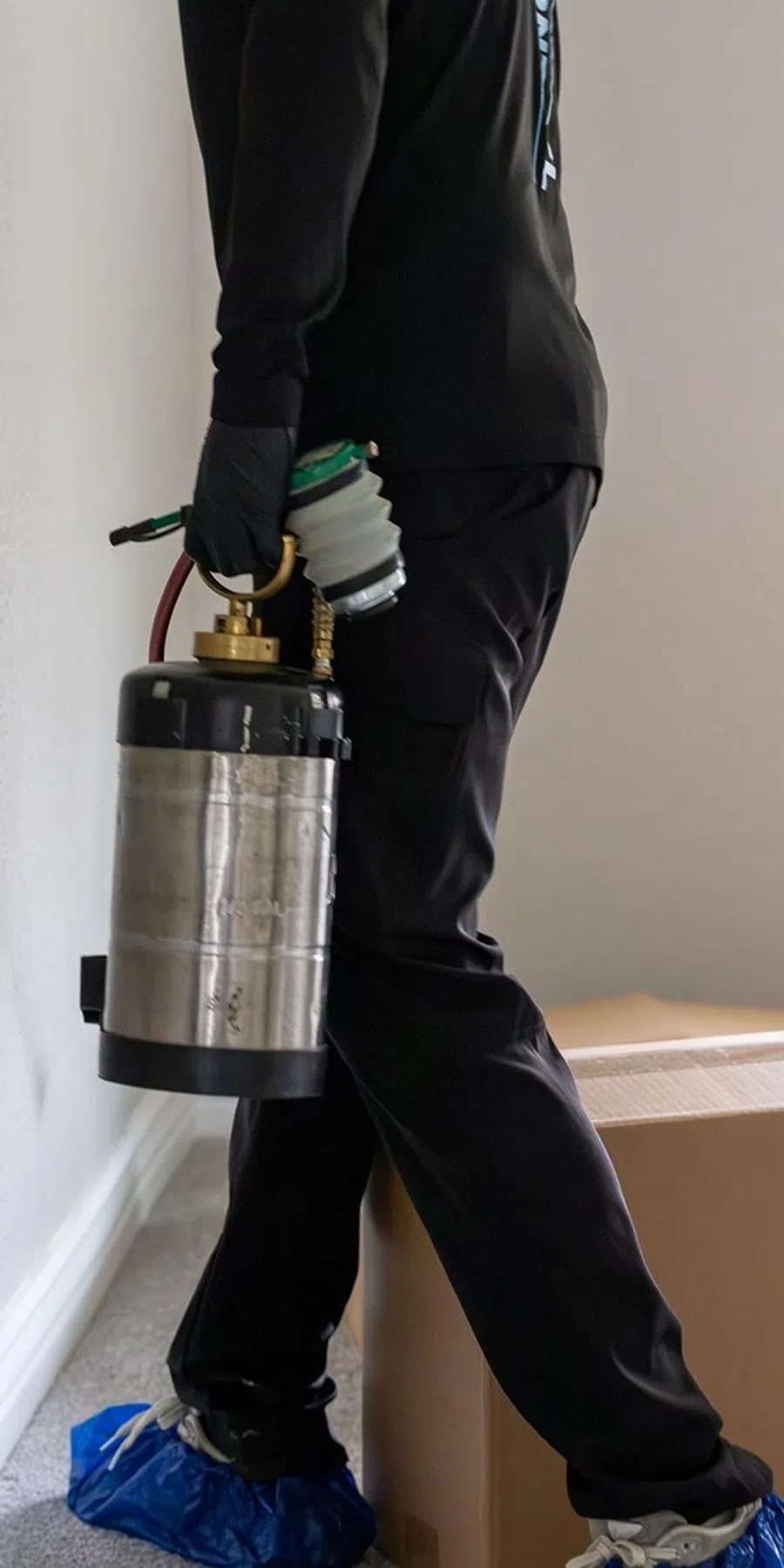 Pest control professional wearing gloves and shoe covers holding a metal sprayer indoors.