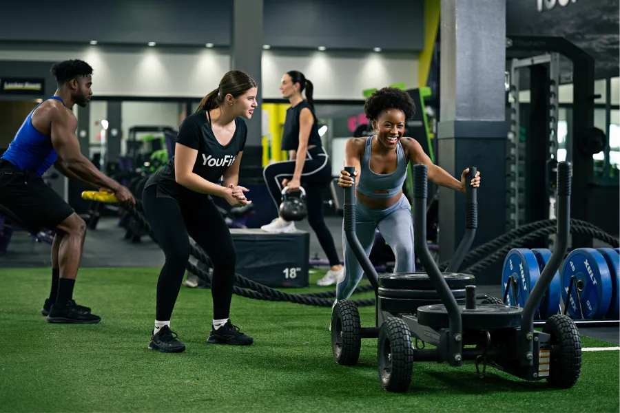 Group of diverse people working out in gym pushing weighted sled and exercising with kettlebells on turf.