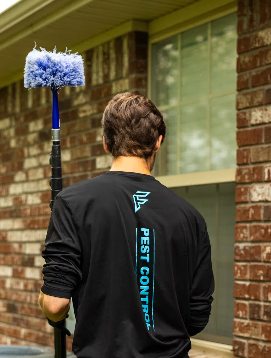 Man wearing pest control shirt holding a blue brush standing outside a brick house near a window.