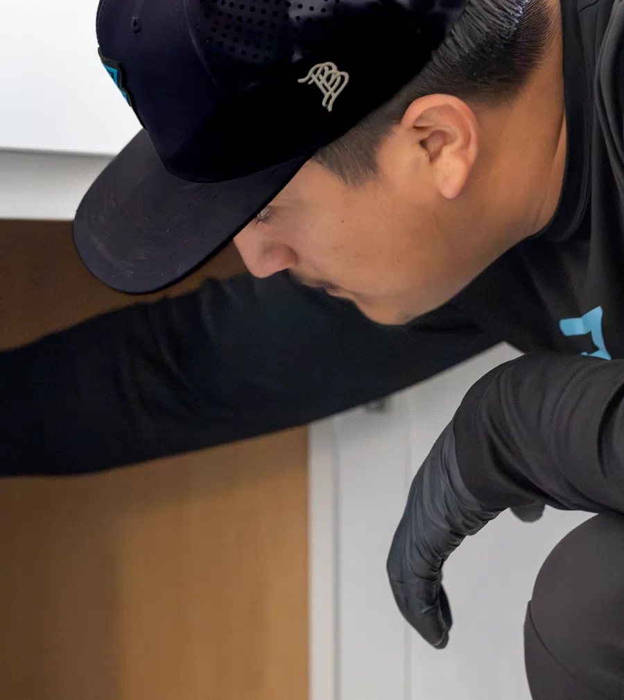 Man wearing black cap and gloves reaches inside wooden cabinet under white countertop in kitchen