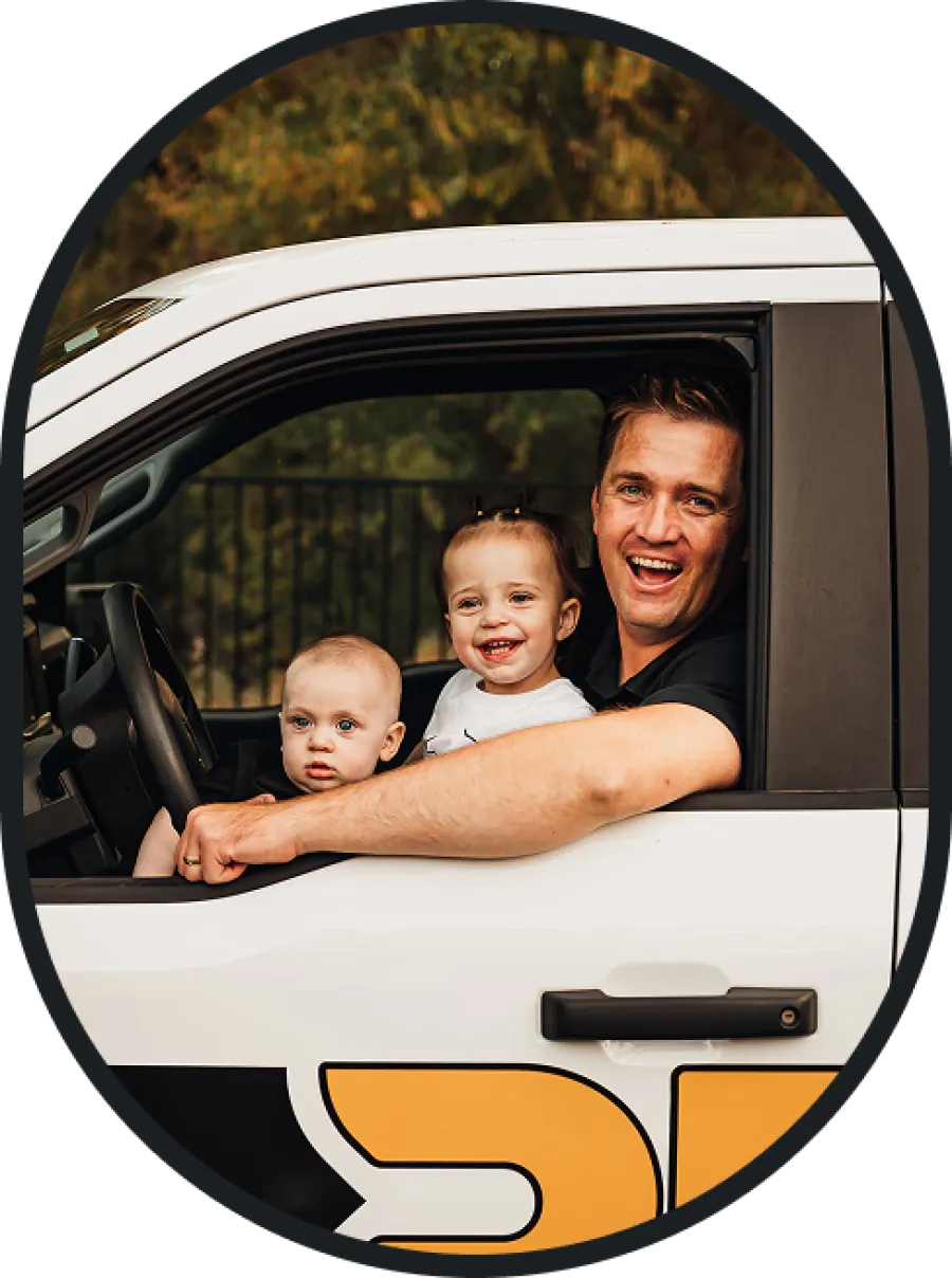 Smiling father and two young children sitting inside a white car with a yellow and black logo on the door.