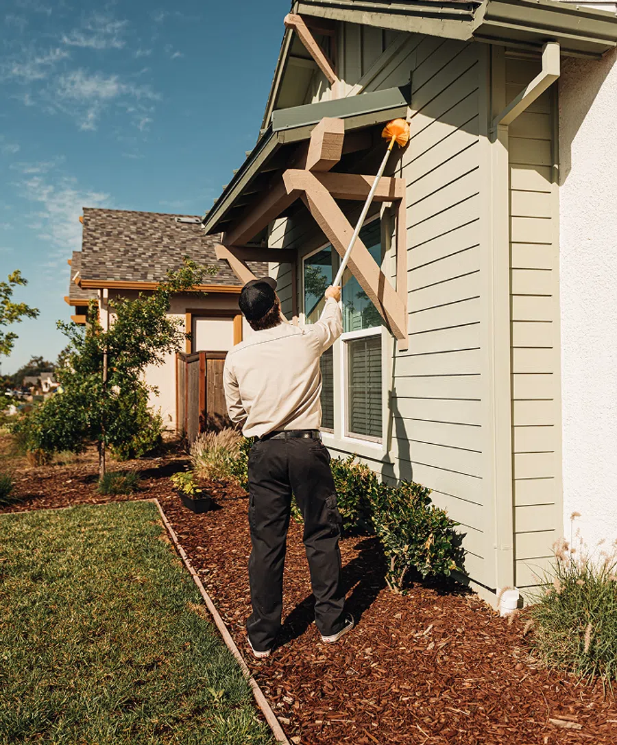 Man cleaning exterior of house with long-handled duster on sunny day in suburban neighborhood