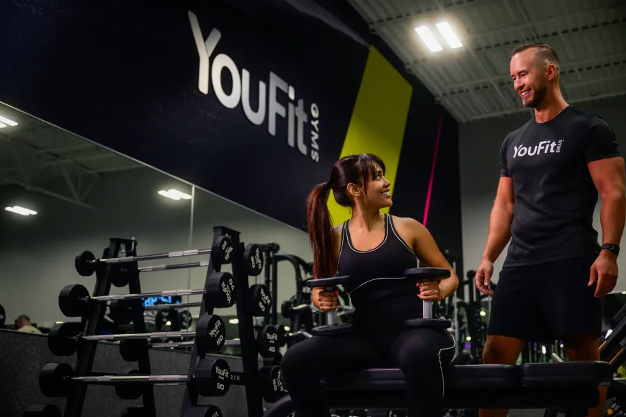Female exercising with dumbbells on bench guided by male trainer at YouFit gym under bright lights.