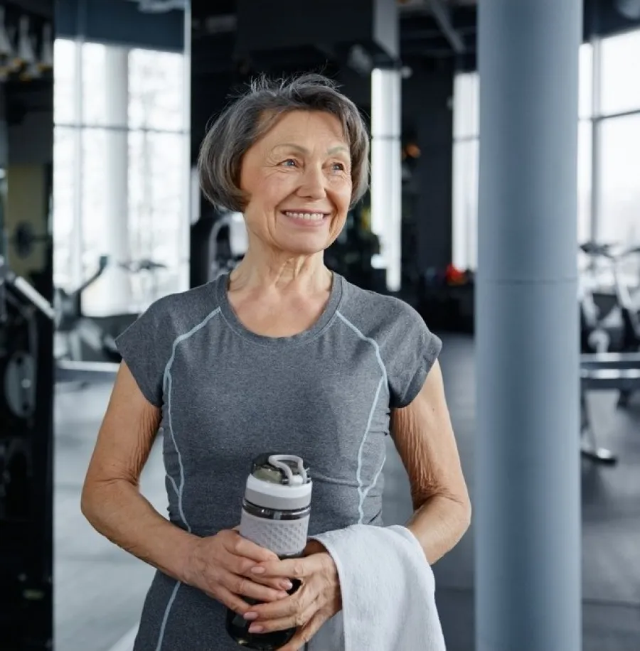 Smiling senior woman in gym attire holding water bottle and towel, standing in modern fitness center.