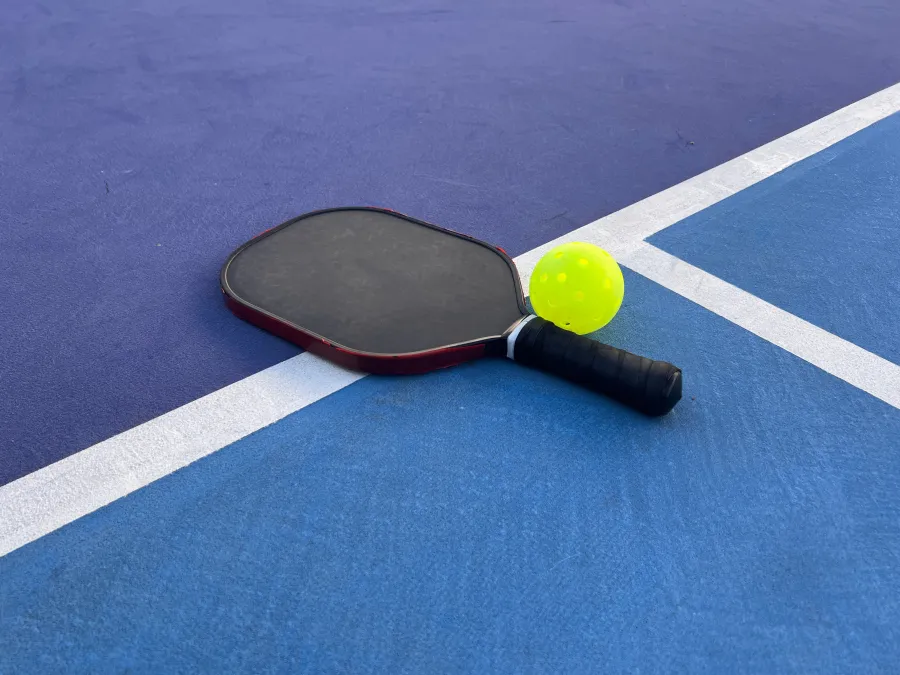 Pickleball paddle and yellow perforated ball on blue and purple court with white lines.