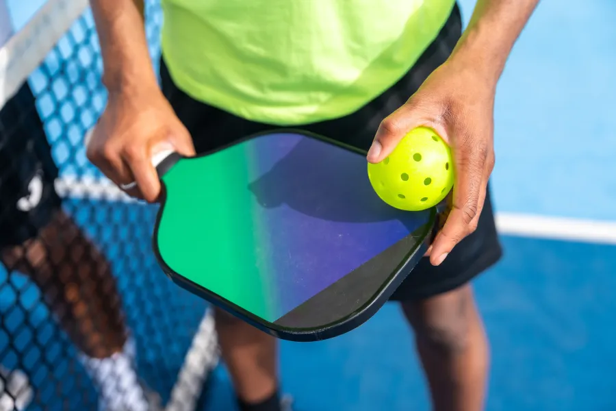 Player holding a green and black pickleball paddle with a yellow perforated pickleball on a blue court.