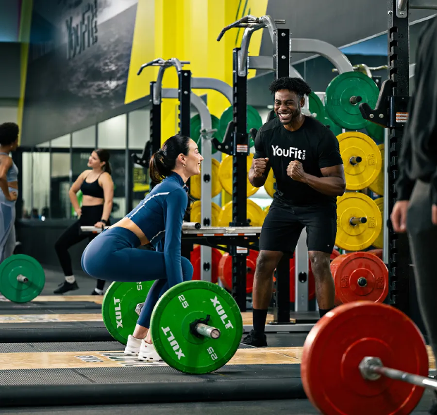 Woman lifting barbell while male trainer cheers her on in vibrant gym with colorful weight plates