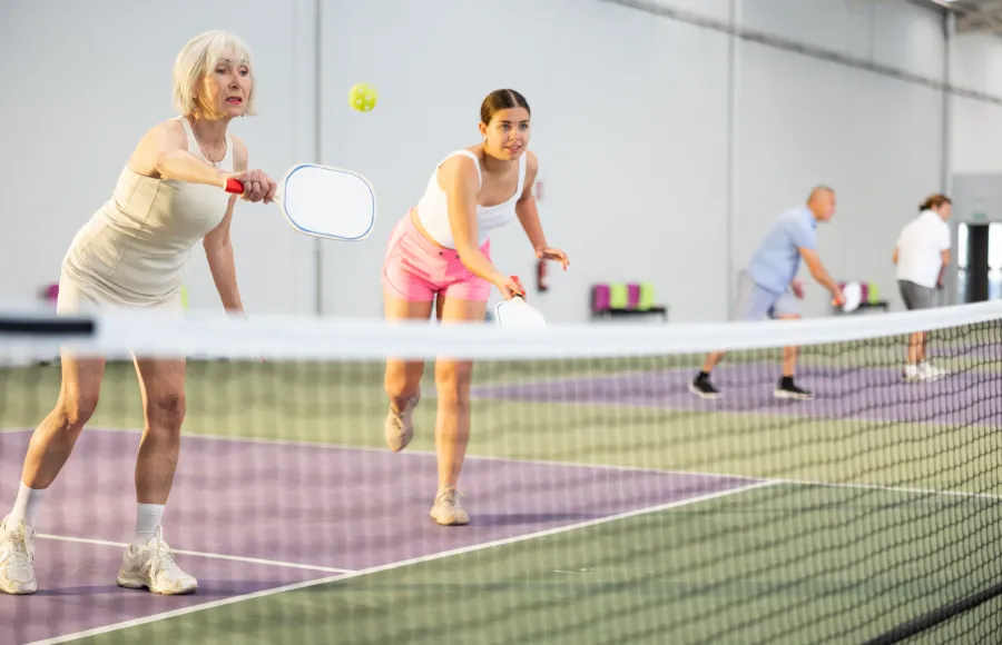 Two women playing pickleball indoors with two men in the background on a purple and green court.