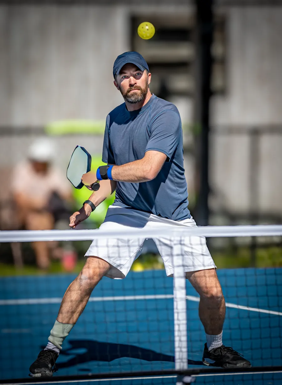 Focused man in blue shirt and cap playing pickleball hitting a ball over the net on an outdoor court.