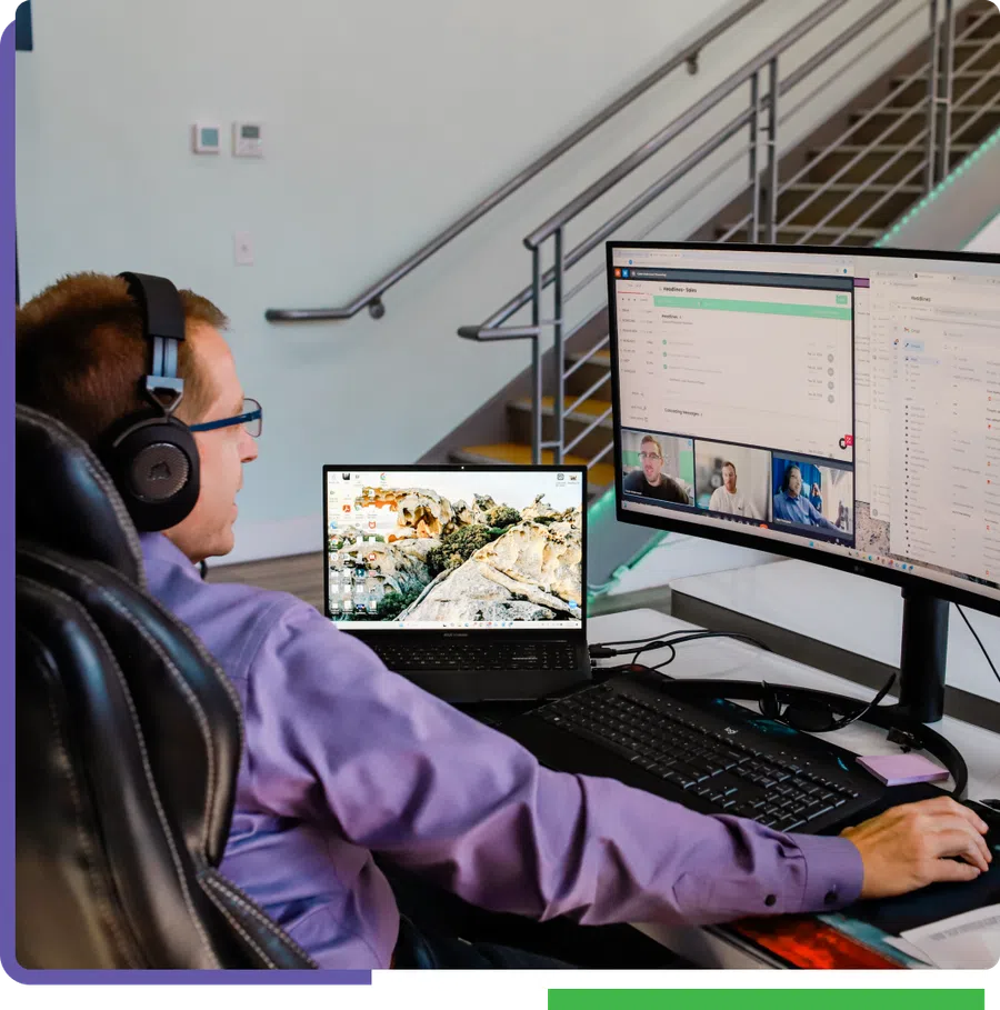 Man wearing headphones working on laptop and large monitor with video call and project management app open.