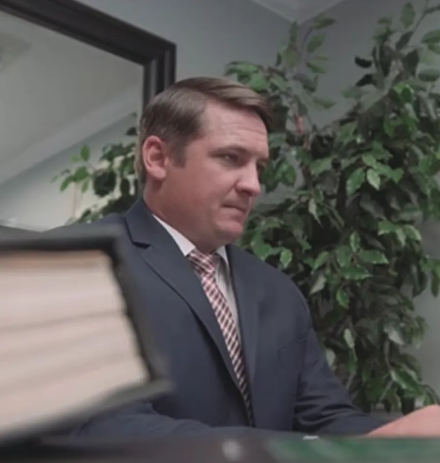 Man in suit and tie sitting at desk with large book and green plant in office setting