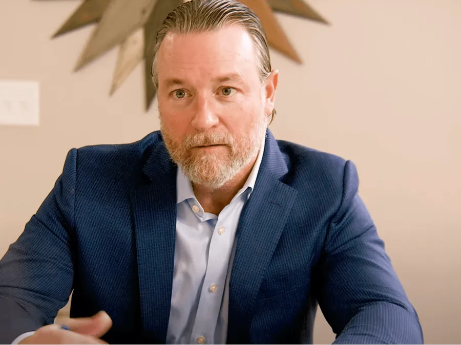Bearded man in a blue blazer and light blue shirt sitting indoors with a neutral background.