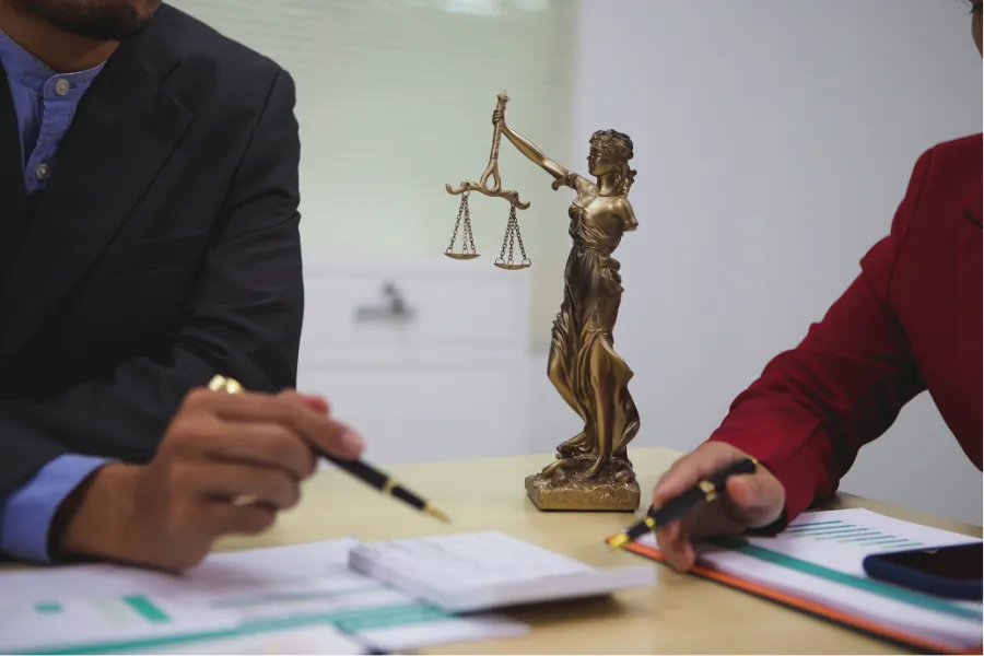 Two professionals discussing documents at a desk with a Lady Justice statue and calculator in view.