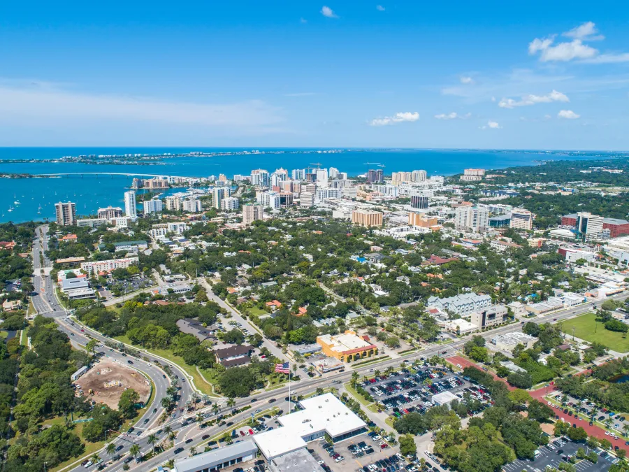 Aerial view of a coastal city with high-rise buildings, green parks, roads, and clear blue water under a bright sky.