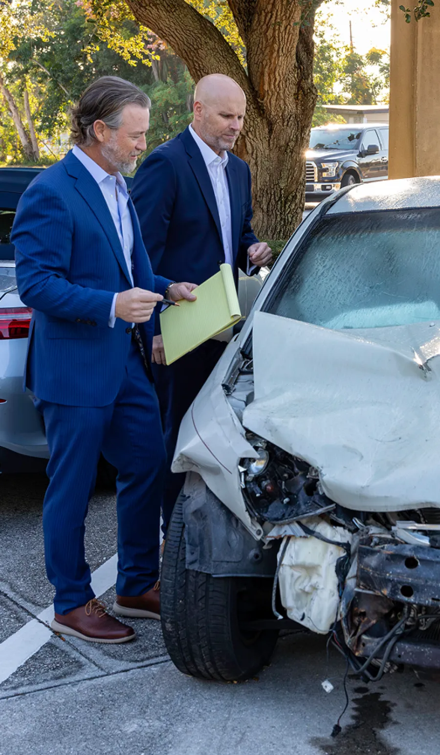 Two men in suits inspecting damaged white car after accident, one holding yellow notepad and pen outdoors.