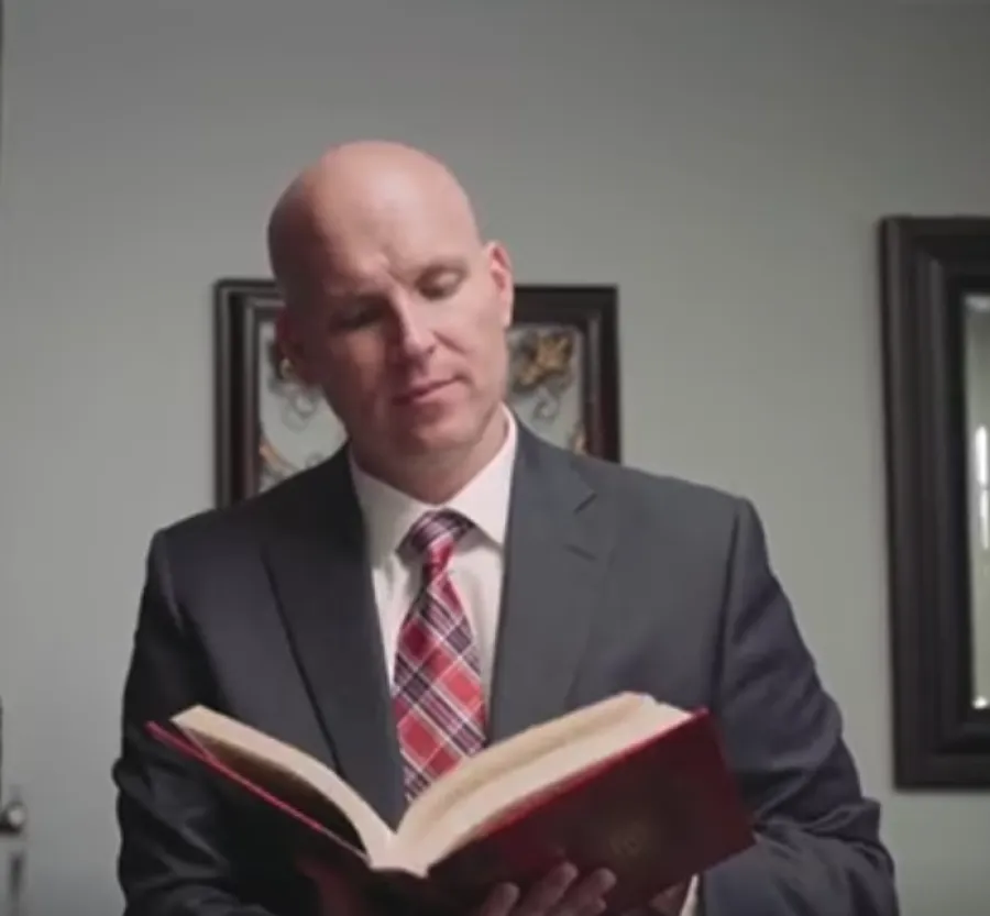 Bald man in suit and red plaid tie reading a book in a room with framed artwork on the wall.