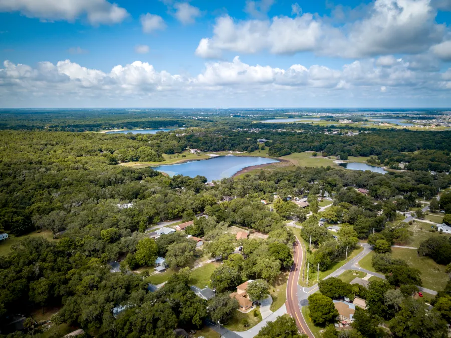 Aerial view of a green suburban neighborhood with winding roads, trees, and lakes under a blue sky with clouds.