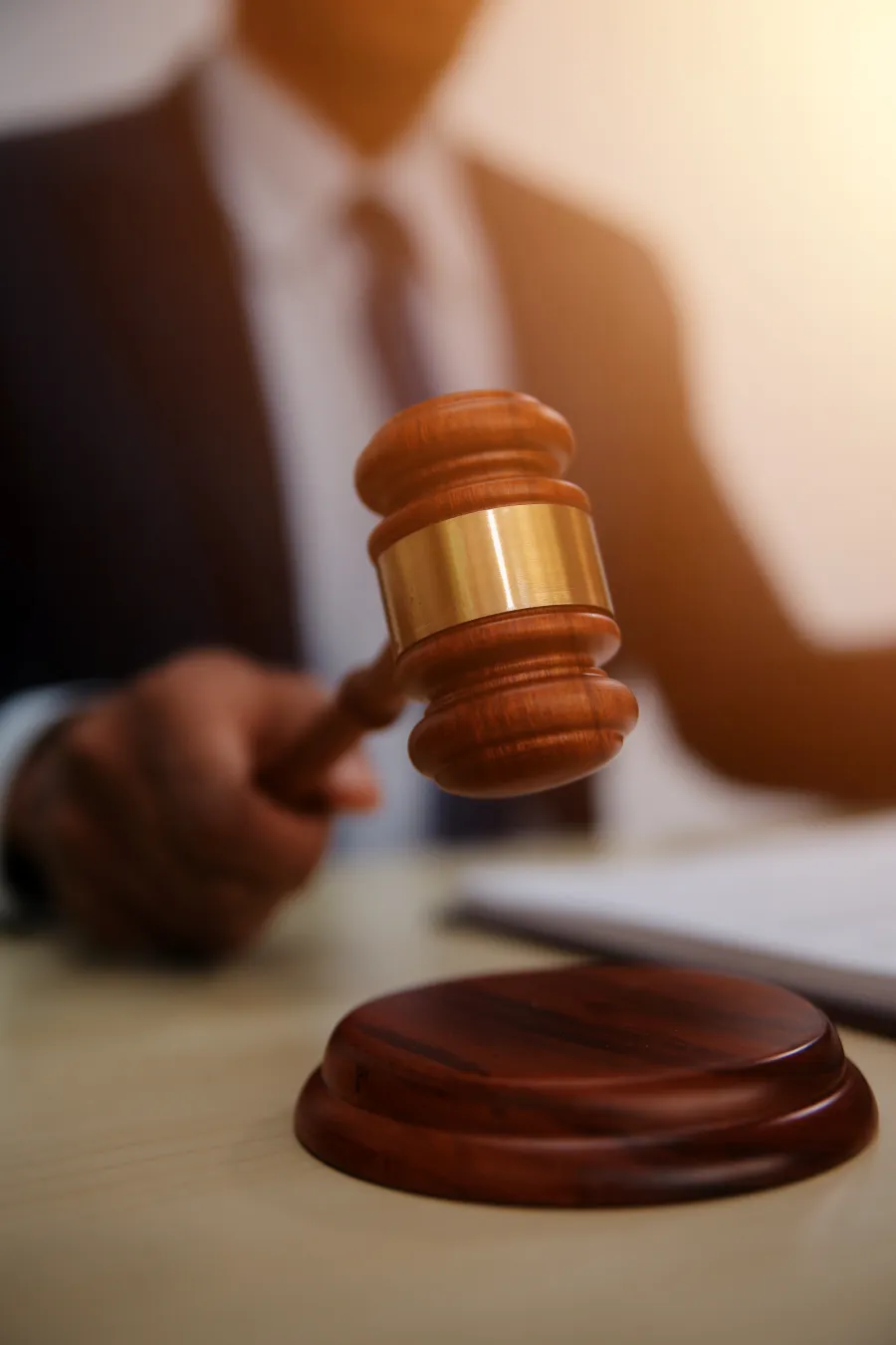 Close-up of a judge in suit striking a wooden gavel on a sound block in courtroom setting.