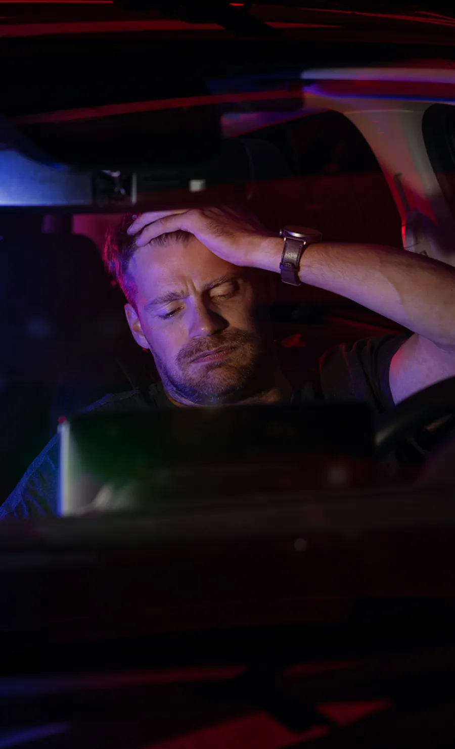 Man sitting inside a car at night, holding his head with a stressed and tired expression under blue and red lights.