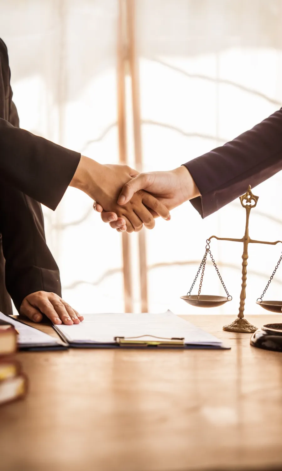 Handshake between two professionals over legal documents with scales of justice on wooden desk.