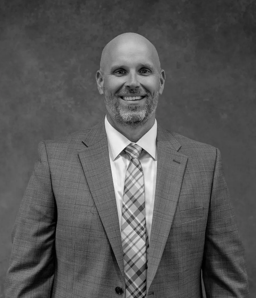 Smiling bald man in a plaid suit jacket and patterned tie against a textured gray backdrop.