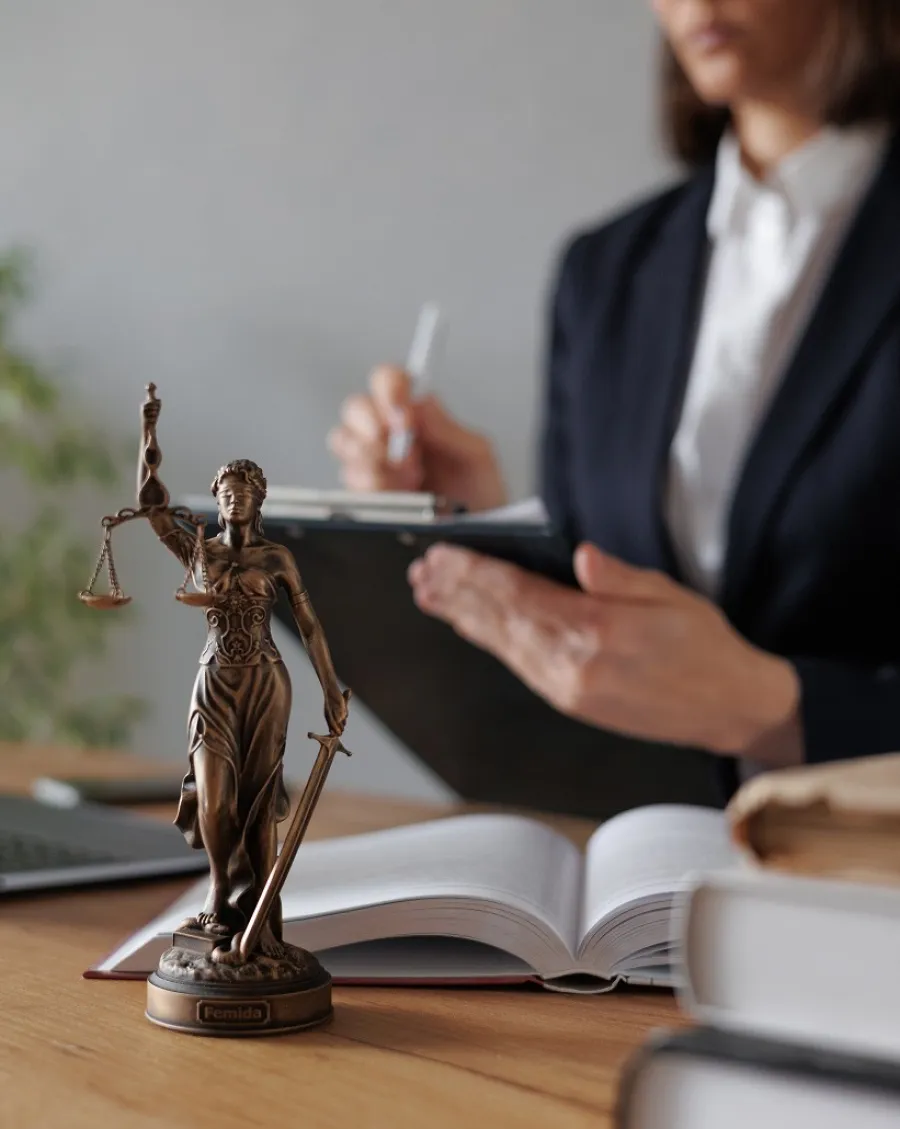 Bronze statue of Lady Justice with scales and sword in front of a lawyer writing on a clipboard over open legal books.