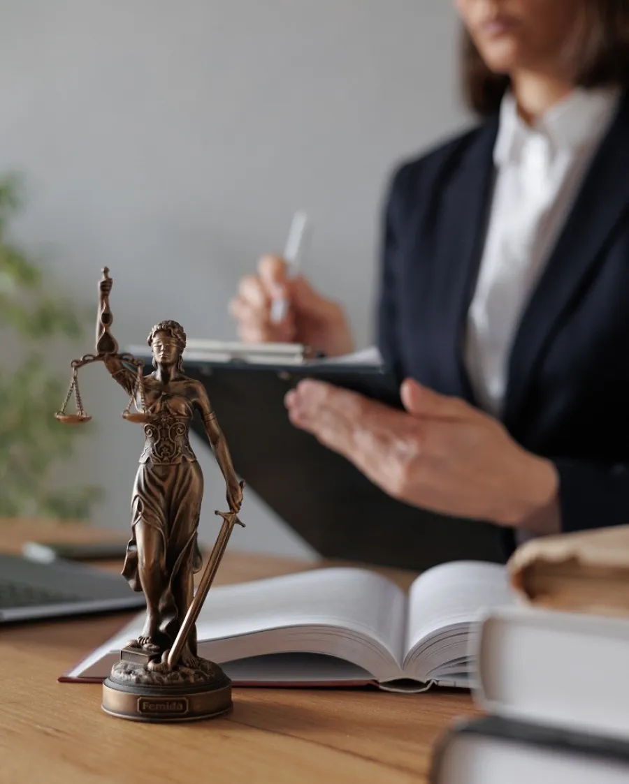 Lady Justice statue in focus with a lawyer writing on a clipboard and legal books on a wooden desk.