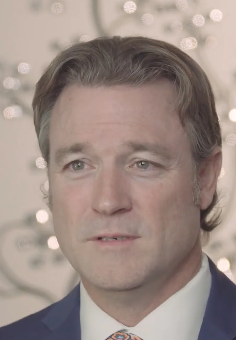 Close-up of a middle-aged man with light brown hair wearing a suit and patterned tie against a blurred background.