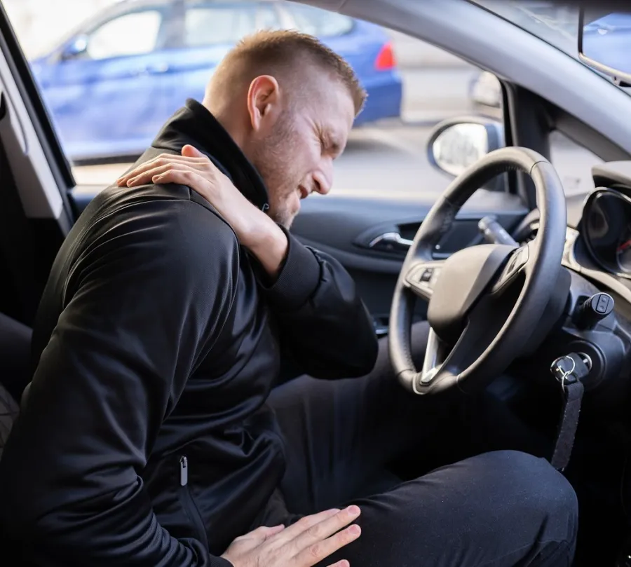 Man sitting in car holding neck in pain, showing discomfort inside the driver's seat of a vehicle.