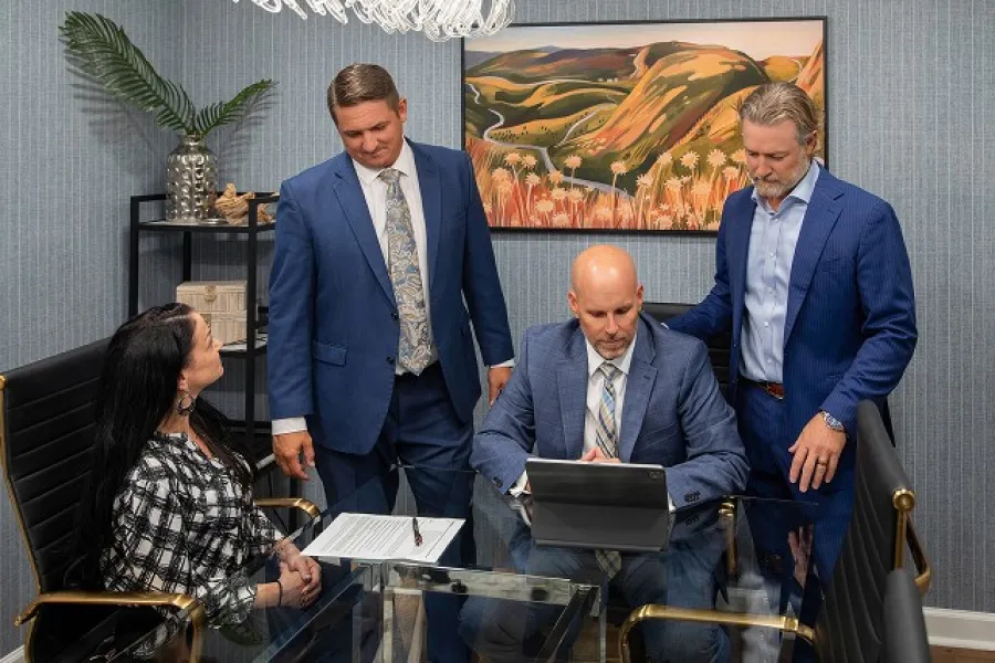 Business team of four professionals in suits discussing work around a glass conference table in modern office.