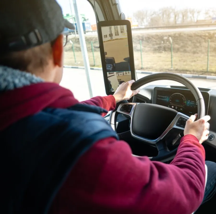 Truck driver wearing a cap and red jacket driving a modern vehicle with digital dashboard and screen in daylight.