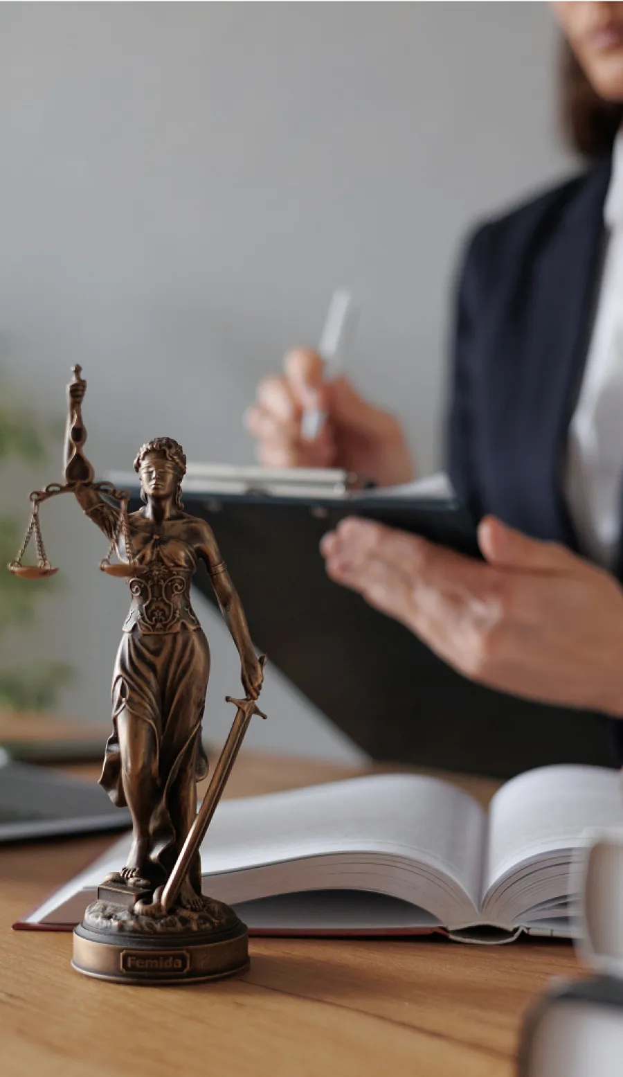 Bronze Lady Justice statue with scales and sword on desk, lawyer working with open book and clipboard in background